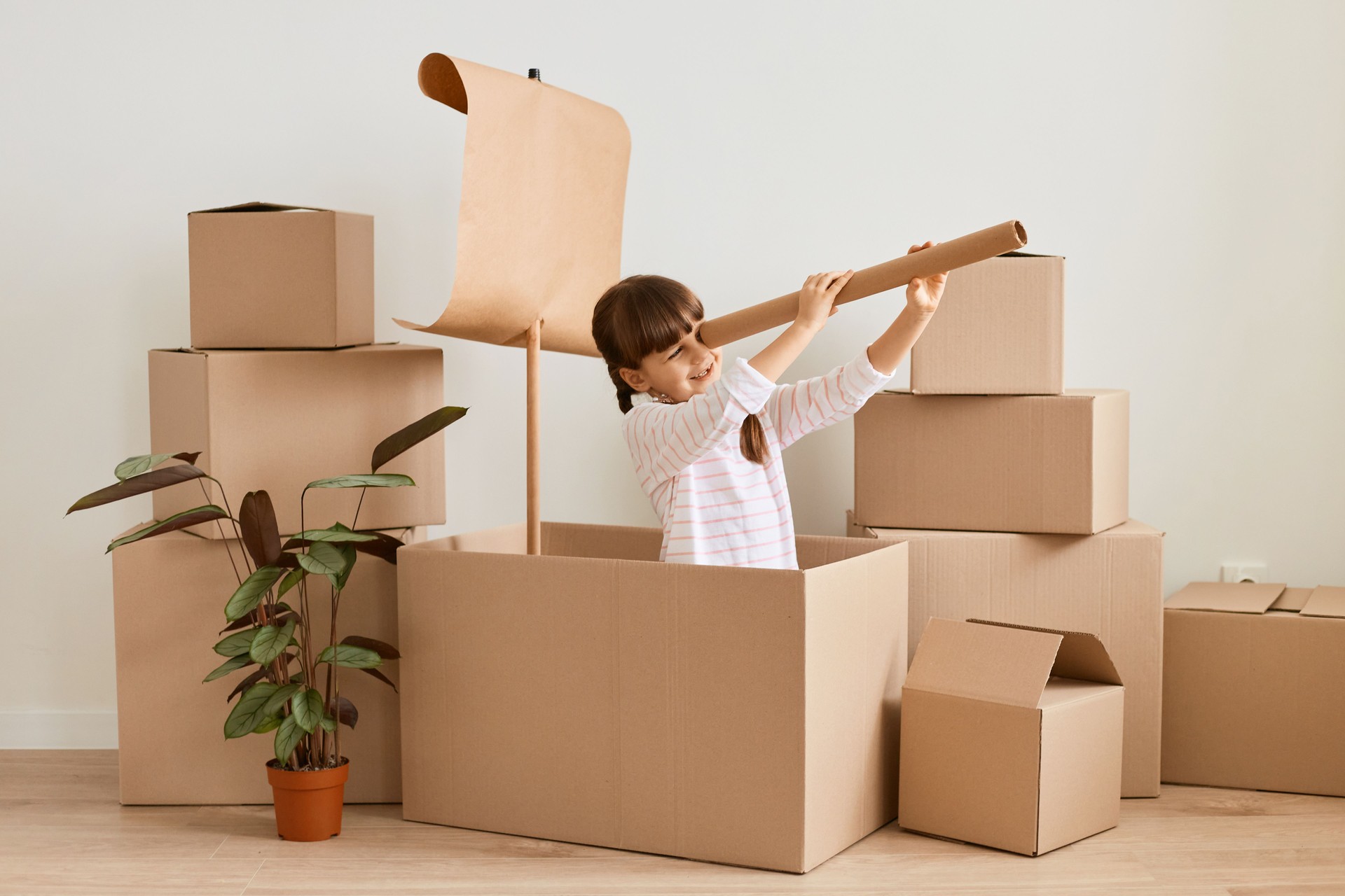 Indoor shot of little girl with dark hair and braids sitting in handmade ship from cardboard boxes and looking in paper spyglass, child playing during relocating.